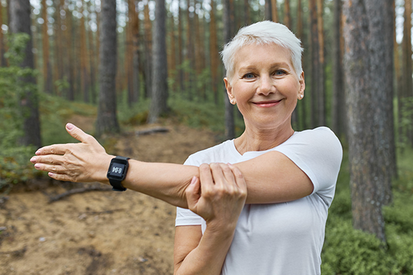 Close up shot of attractive short haired retired female wearing whit t-shirt and smart watch on her wrist to track progress during running, stretching arm, preparing body for cardio exercise